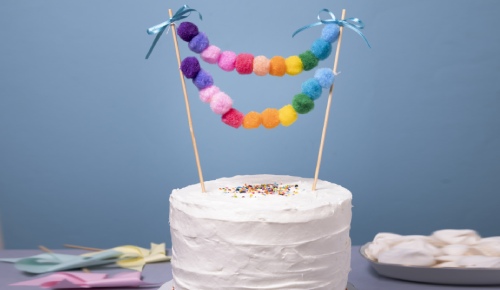 A simple white-frosted cake adorned with a colorful garland of fluffy pom-poms, held between two wooden sticks