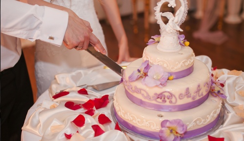 A couple in weeding dress cutting into a multi-tiered wedding cake adorned with purple strip and fresh flowers