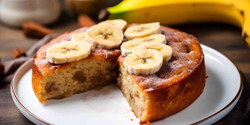 A round banana cake on a white plate with a slice removed. The cake is topped with fresh banana slices.