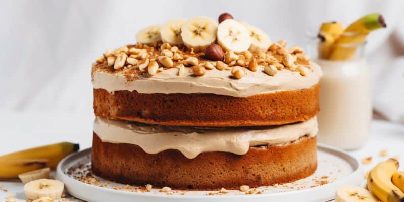 A two-layer banana cake with creamy frosting is topped with sliced bananas and chopped nuts. 