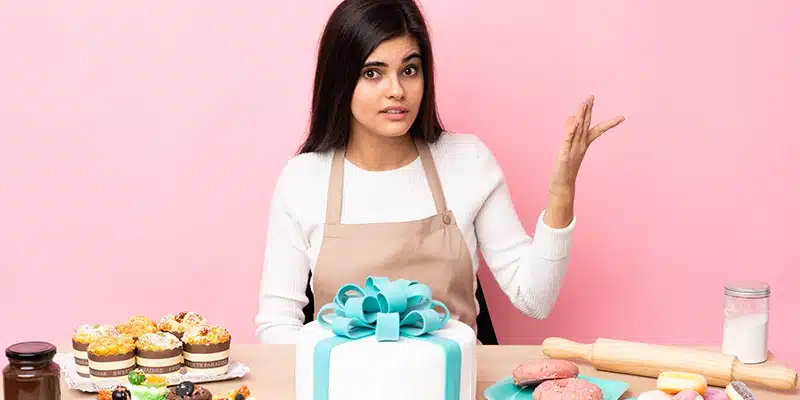 A baker with a white apron, showcasing various baked goods including custom cakes, pink frosted donuts, and cupcakes. Factors influencing the price, such as ingredients and decoration, are visible.