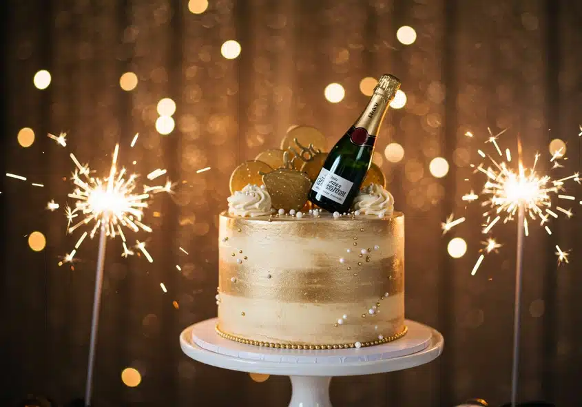 A glamorous New Year's Eve champagne cake decorated with a gold theme, edible pearls, frosting swirls, golden toppers, and a mini champagne bottle, with sparklers and a sparkling backdrop, showcasing "New Year's Eve Cake Celebrations: Champagne Cake."