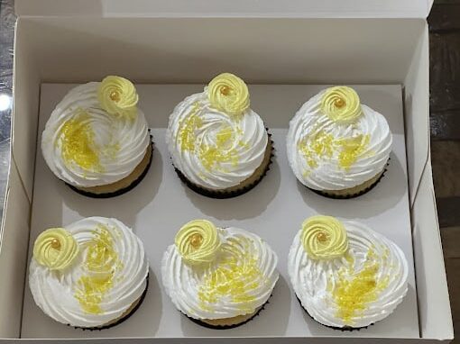 Six lemon cupcakes in a white box, each topped with white frosting, yellow swirls, and sprinkled yellow sugar crystals.