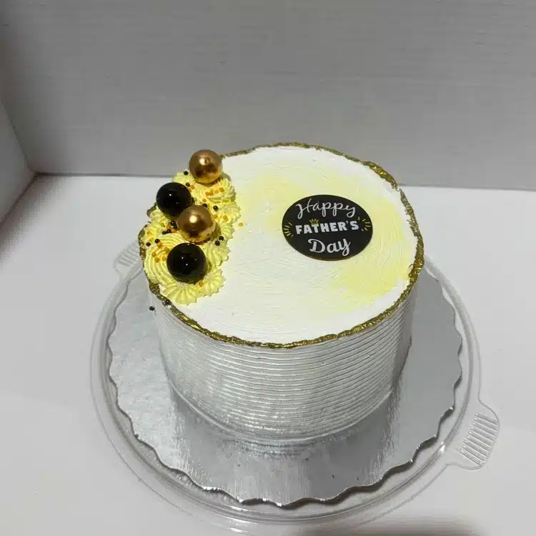 A classic Father’s Day cake with white frosting, gold and black edible balls, yellow cream accents, and a “Happy Father’s Day” topper.