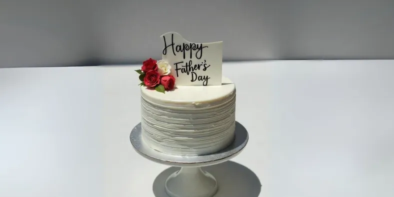 A beautiful Father’s Day cake with white frosting, red roses, and a topper reading “Happy Father’s Day” on a white cake stand.