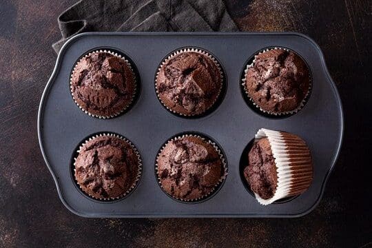 Six freshly baked chocolate fudge cupcakes in a dark muffin tray, with one cupcake partially unwrapped to show texture.
