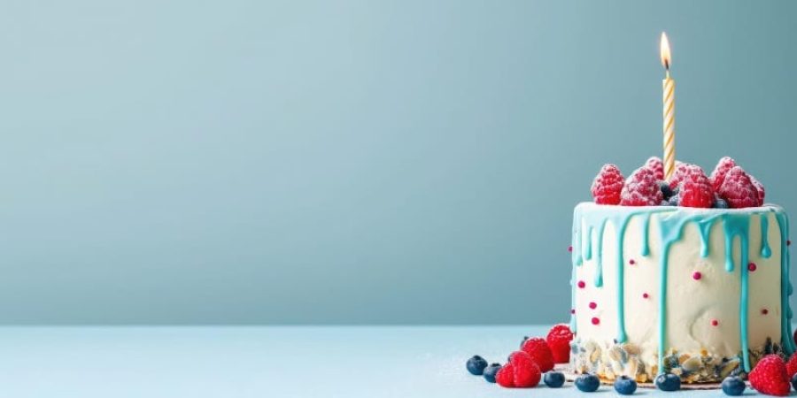 A single lit candle tops a small custom birthday cake adorned with dripping blue icing, fresh raspberries and nuts and berries.