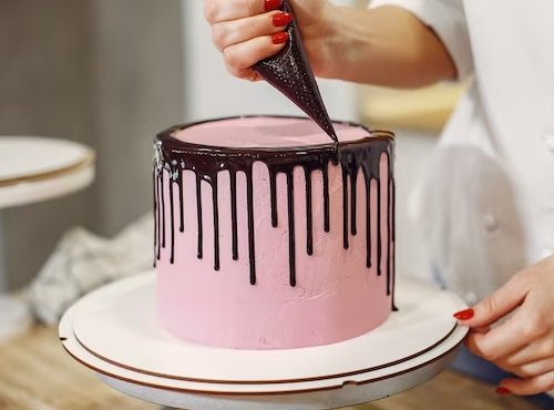 A baker expertly applies a chocolate ganache drip to a beautifully frosted pink cake